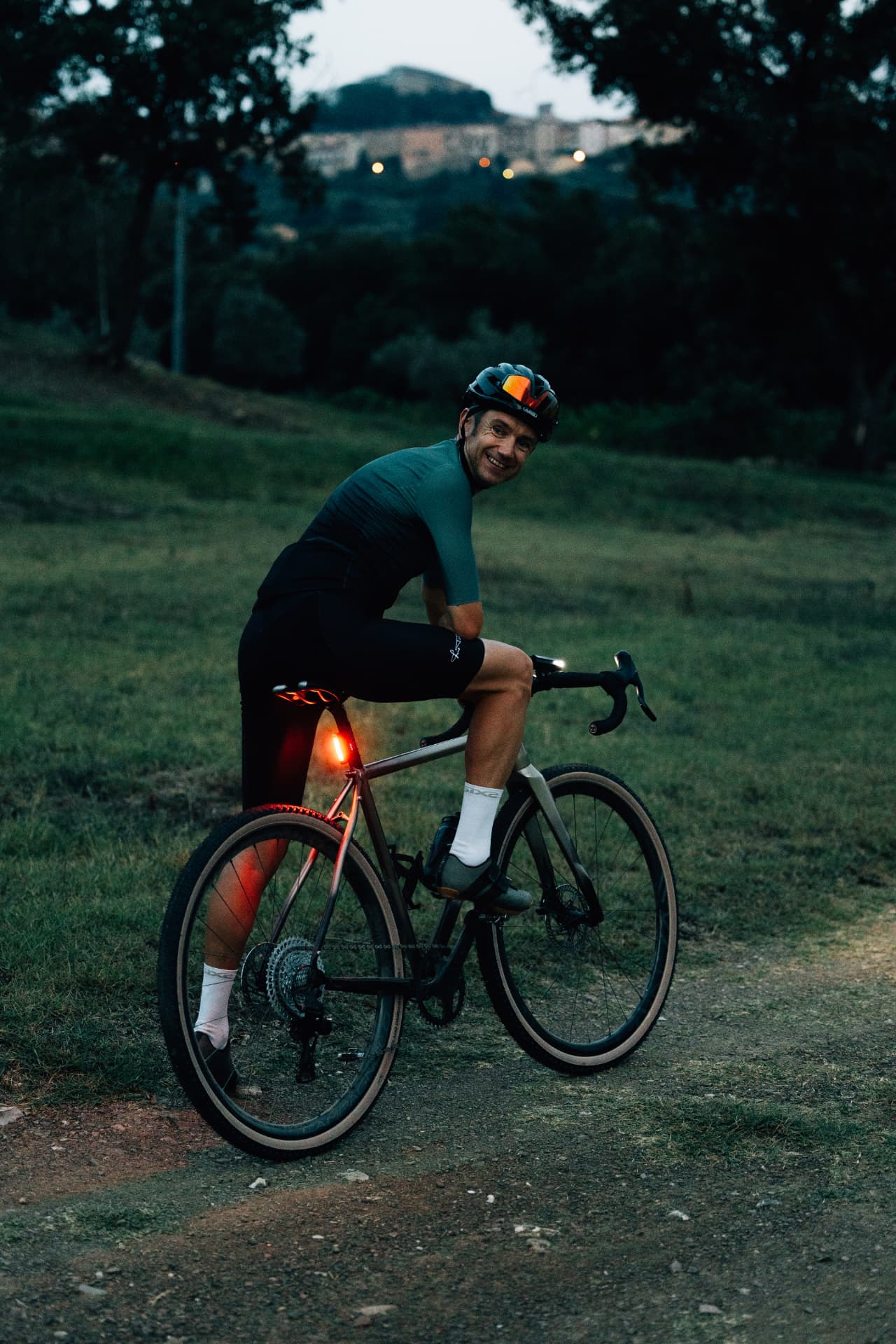 Cyclist with the Tommasini Fire at dusk, village lights on the hill behind