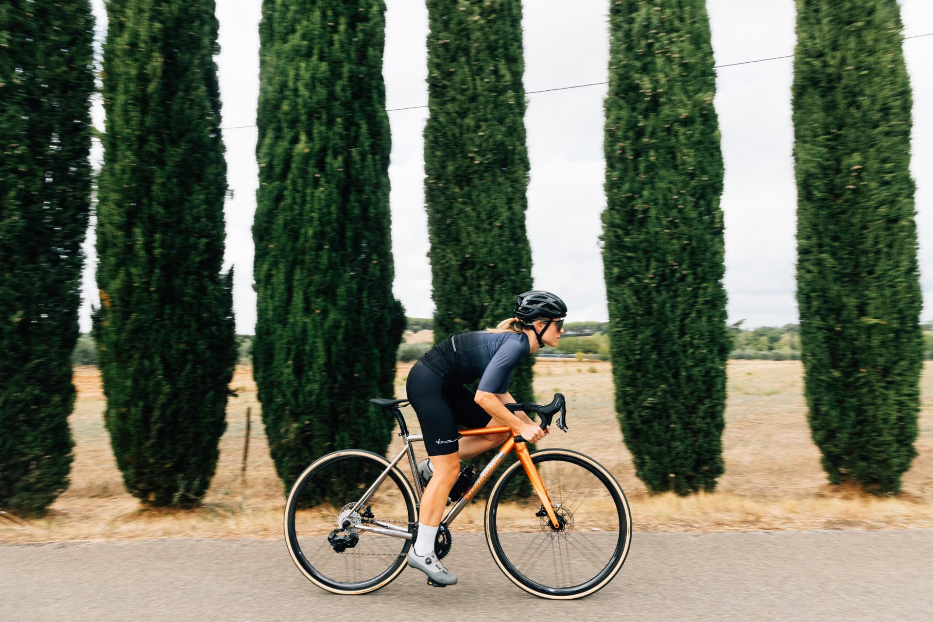 Cyclist riding the titanium and orange Mach Ti road bike along a Tuscan cypress-lined road